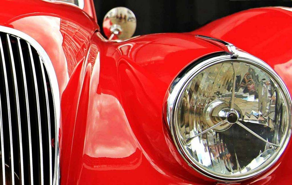A close up image of an old Jaguar sports car viewing the fron headlight and a portion of the front radiator grill. The car body is painted in a luxurious red and is highly polished, waxed and shiny.