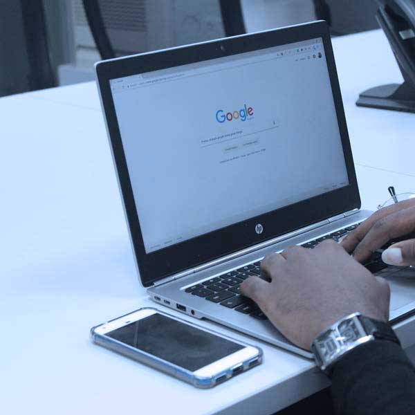 showing 2 male hands typing on a notebook computer keyboard. The computers screen is showing the home page for Google, ready to type in a search. The notebook computer is sitting on a large white desk and there is a momile phone next to the laptop.