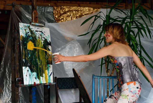 A female artist throwing yellow paint onto a canvas on an easel. the paint is making a splash on the canvas as the paint leaves a cup being held by the woman at arms length as she throws.